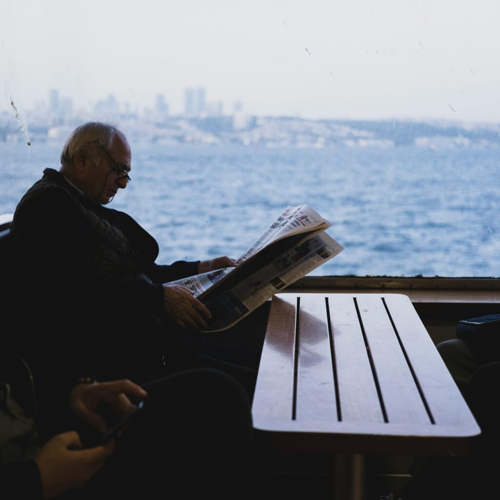 Senior man enjoying a quiet moment reading a newspaper on a ferry with a cityscape view.