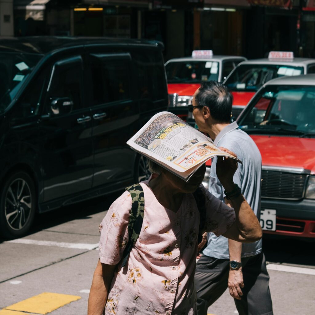 Two people walk through an urban street, one using a newspaper for shade.