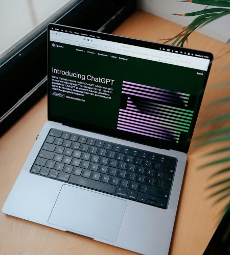 A sleek laptop on a wooden desk showing the ChatGPT homepage by a window.